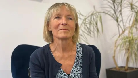 Dr Sian Wareing-Jones sat on a blue arm chair. She is wearing a blue cardigan and blue floral dress. The walls around her are white and there is a plant behind her. 