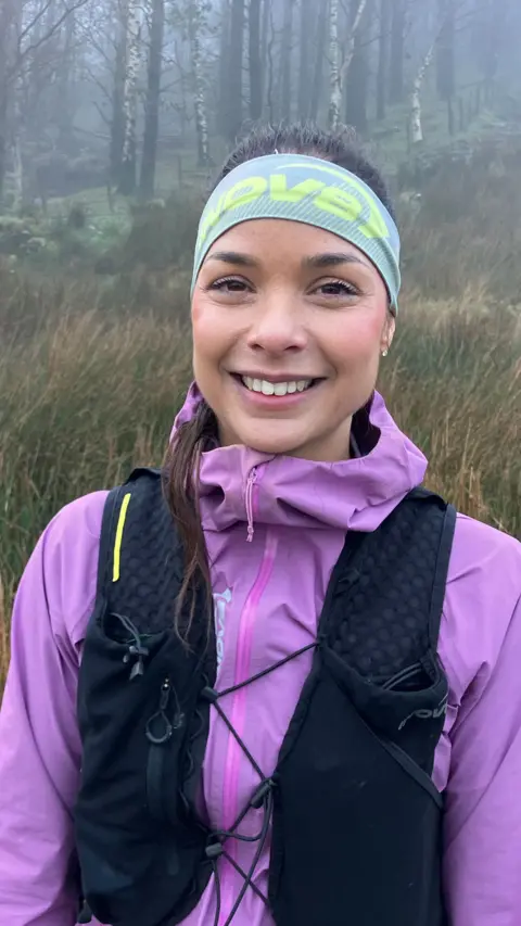 Sarah Perry. She is smiling and wearing a black running bag over a purple jacket. Her brown hair is tied back and she is wearing a headband. Behind her are trees and grassy bushes.