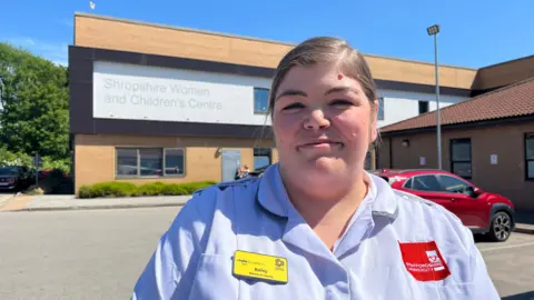 Bailey pictured standing outside the Shropshire Women and Children's Centre at the Princess Royal Hospital in Telford - it's a large modern brick building with 'Shropshire Women and Children's Centre' written on the outside. Bailey is wearing a pale blue nurse's tunic, with a red University of Staffordshire logo on the left side, and a yellow NHS badge that reads 'Hello my name is Bailey' on the right. The sky is bright blue and clear, and there's lots of sunshine. 