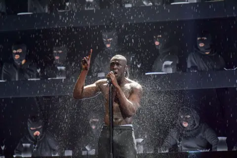 Getty Images Stormzy performs Blinded by Your Grace, Pt. 2 on stage at The Brit Awards in 2018. He's stood singing into a microphone with his left hand on the mic and his right hand in the air with his index finger pointing to the sky. He has black jogging bottoms on and is topless. There's water falling from the ceiling on to him and behind him there's a choir made up of people dressed all in black including black balaclavas.