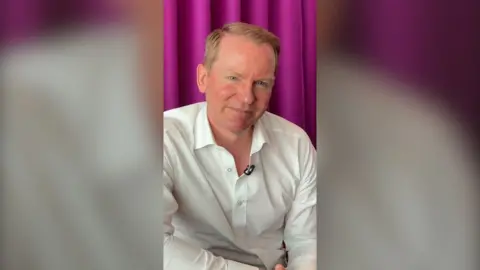 A man, Charlie Nunn, wears a white shirt. He has short red hair and sits in front of a purple curtain.