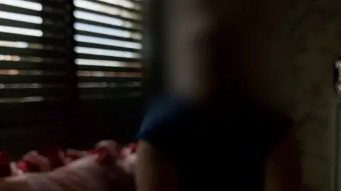 Blinds are open on the left of the photo in a dark room. A woman whose face is not visible is wearing blue on the right.