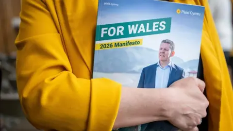 A woman in a yellow jacket holding a copy of the Plaid Cymru manifesto, which reads "for Wales" in capital letters and "2026 manifesto". The cover of the manifesto shows Rhun ap Iorwerth looking off into the distance.