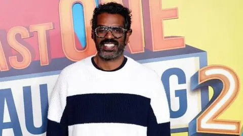 Romesh Ranganathan is seen during the photocall for Prime Video's Last One Laughing. He is wearing a black and white jumper and smiling and the logo for the programme is behind him.