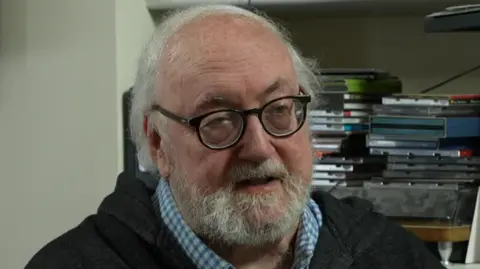 Martin Shipton, journalist, seated and wearing a checked shirt and a gilet, at his home in Cardiff