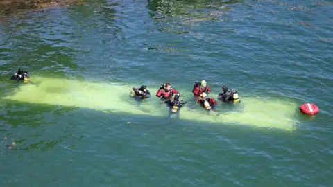 Reuters The roof of the bus is visible under the river. Six people in wetsuits are sat atop it, and one more person is in the water on the left of the frame.