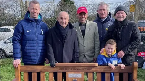 A group, including Micky Hazard, Ossie Ardiles and Bryan Hughes, stand behind a memory bench