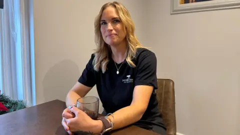 Martell Blackford sat at a table with a coffee in a glass mug. She has blonde air and is smiling. She is wearing a necklace and a black t-shirt that says 'Jacob's Table'. She is wearing a watch and some bracelets. She has a tattoo on her left hand. 