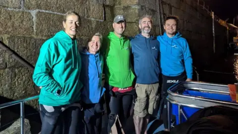 Five swimmers standing on steps below a harbour, next to a boat