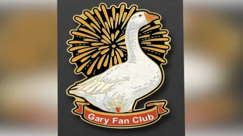 Chrissie Fadipe/Linda Batten A design of Gary the Goose with an exploding orange firework behind him. The goose is white with an orange beak. A banner at the bottom says "Gary Fan Club" conceals the goose's feet.