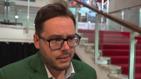  Nick Hartley at Newcastle Civic Centre. He has brown hair and a beard. He is wearing glasses and a green suit.