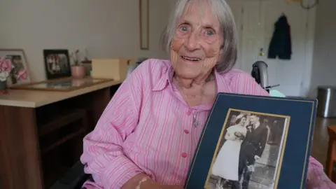 Rita Harvey / Royal Star & Garter an older woman wearing a pink striped shirt sits holding a black and white photograph of her wedding day