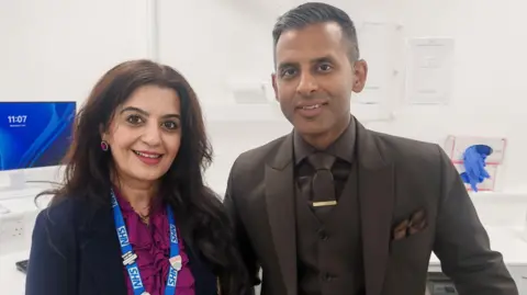 A woman and a man standing in a clinic. The woman is wearing a purple top and an NHS lanyard. The man is in a brown suit. There is a computer screen behind him.