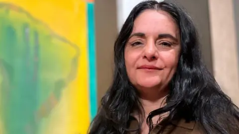 Emma Baugh/BBC Habda Rashid is standing on the right. She has long black hair and is wearing a khaki jumper. Behind her, and on the left, is one of Frank Bowling's paintings which is of Africa. 