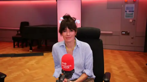 Lena Headey has dark tied-back hair and is wearing a light blue shirt. She is sat in front of a red microphone.