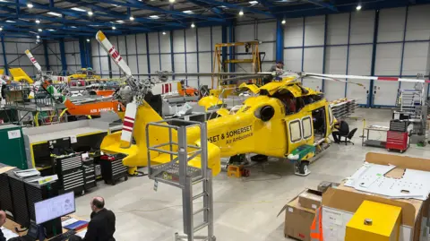 A zoomed out view of yellow air ambulance helicopter, with a man working on its nose. It is in a warehouse-style building, with other air ambulances visible in the background behind shelving and machinery. The helicopter has its door open and there are tools and a small stool on the floor of it. 