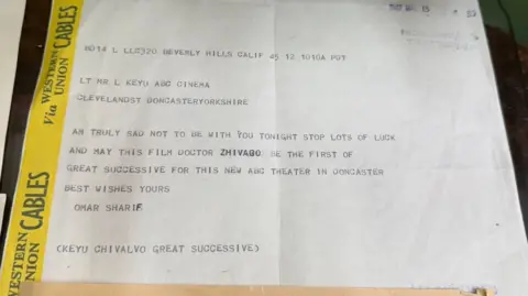 BBC/Grace McGrory A piece of paper with a few creases in it. It contains a printed message which is signed "Omar Sharif". There is a yellow border which runs down the left-hand side.