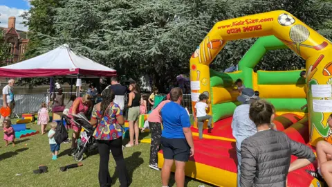 Bedford Borough Council People taking part in events at the River Festival, there is a bouncy inflatable, and a gazebo. Trees are also behind the structures, a building to the left and an area of river. 