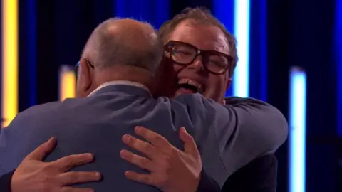 BBC/Hungry McBear Alan Carr hugging Harry Bowden. Alan's face is visible and he is smiling with his eyes close, while Harry Bowden is seen from behind.