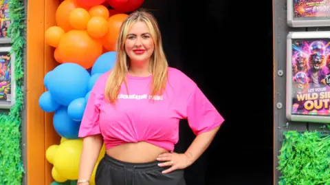 A woman stands in the doorway of a venue decorated with a large rainbow balloon arch, wearing a bright pink cropped T‑shirt and black shorts