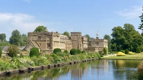 Marika Reinholds Forde Abbey as it looks today - a grand stone mansion with crenelated parapets stands next to a large rectangular lake. The building and herbaceous flower borders are reflected in the water.