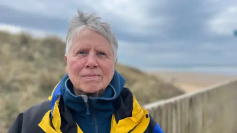 A picture of Sue stood in front of the beach and sea. She is wearing blue and yellow coloured clothing.