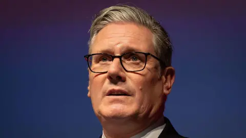 Sir Keir stood at a lectern at Welsh Labour conference. He is wearing a black suit, white shirt and black glasses.