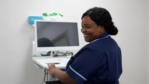 A nurse in a navy uniform operates a computer workstation inside a clinical room, with medical equipment and cables visible on the unit. The nurse is smiling.