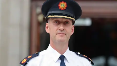 PA Media Justin Kelly looks into the camera. He is wearing a white shirt and black tie. He has shoulder badges with red and yellow designs on a black background. He is wearing a dark hat with gold trim and the Garda Síochána badge.