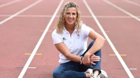 Sally Gunnell, Olympic Gold Medalist poses for a portrait as the next generation of London young athletes are inspired by Team GB performances in Tokyo at London Marathon Community Track on August 03, 2021 in London, England.  She wears a white polo shirt, blue jeans and white trainers.