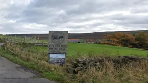 Google A wooden sign next to a road leading down to several buildings nestled in the countryside. The sign reads "The Yorkshire Cycle Hub" and advertises a cafe and other facilities.