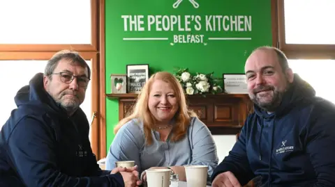 Michael Cooper Damian McNairney, with glasses, (left), Naomi Long (centre), with red hair, and Councillor Paul McCusker (right) in front of green sign of The People's Kitchen Belfast