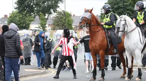 PA Media Man in balaclava stares as police officers on horses