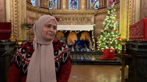 Romana Kazmi, wearing a headscarf and festive red cardigan, in the chapel at Great Ormond Street Hospital, with a lit Christmas tree and nativity scene in the background under the word 'Alleluia'.