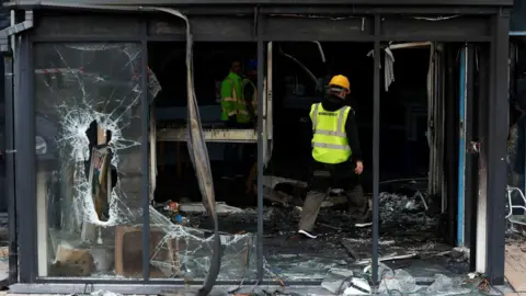 A man wearing a yellow hard hat, a green hi-vis jacket and black workwear trousers, walks through rubble at an entryway to Larne Leisure Centre. There is broken glass door to the left of him. 