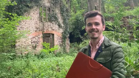 A man standing in the woods with the ruins of an old mill behind him. The ruin is made of bricks and is overgrown with ivy. The man is carrying a red briefcase.