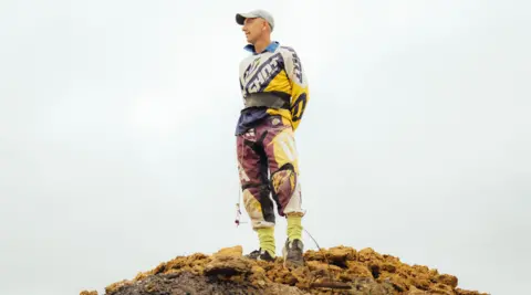 Harry George Hall A man stands on top of a mucky hill dressed in racing leathers