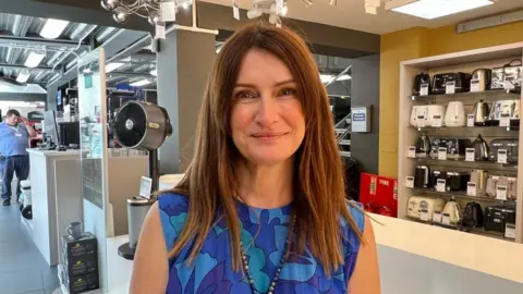 Carla Martin has brown hair and is smiling at the camera. She is wearing a sleeveless blue dress with flower patterns in different shades of blue. She stands in front of a shelf which is empty, except for one remaining fan. She has a necklace of dark and light pearls. 