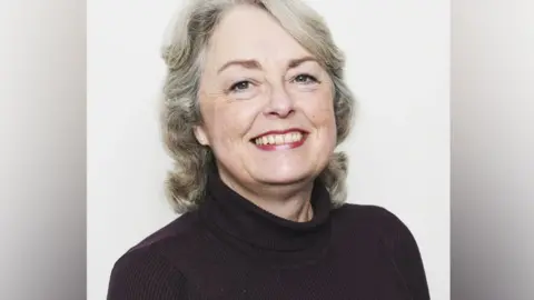 Jacqui is smiling at the camera in a posed photo. She is wearing a dark polo neck jumper and has shoulder length grey hair