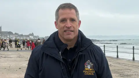 James has grey hair and is wearing a dark blue jacket with the logo of Penzance Council on it. He is standing on the prom with the sea, town, and some people behind him. 