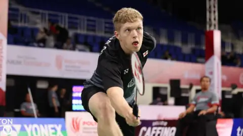 Badminton England Rob Donald, wearing black shorts and top, stretching out to hit the shuttlecock during a game of badminton. People watch the rally from blue seats in the stadium.