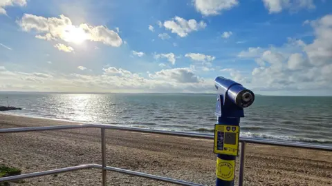 stacey johns A beachside telescope overlooks yellow sands and a calm-looking sea with sun reflecting off the water. 