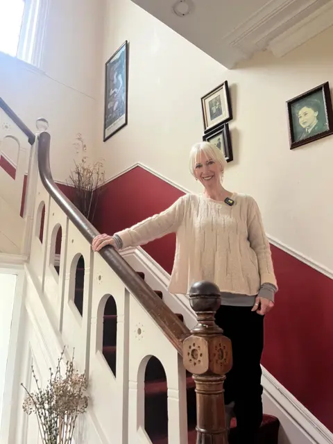A woman with short white hair smiles as she stands on a grand-looking staircase with a dark-wood bannister and white balluster with decorate arch design. She is wearing a white jumper and dark trousers. The carpet is burgundy and the walls are painting in burgundy and white. Black-and-white photos are framed on the wall. Light streams in from a window high above. 