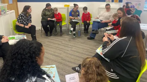 About 8 pairs of children with a parent. They are sat in a circle and all pointing at books or other learning materials together. 