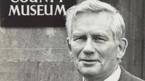 A black and white photograph of Roger, who is dressed in a suit and wearing a stripy tie. He is standing in front of a sign that says Armagh County Museum.