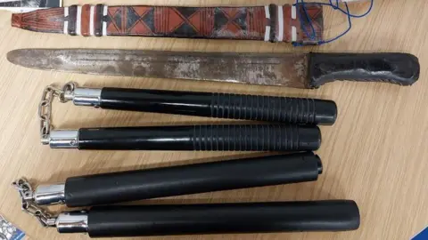Cambridgeshire Constabulary A long knife is on a wooden table beside two nunchucks which are black and silver.