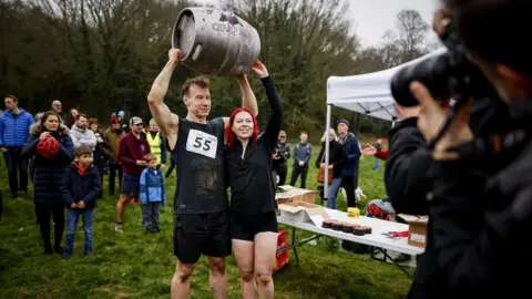 PA Media Two people stand triumphantly. They have just won a race and are being photographed holding a barrel of beer. 