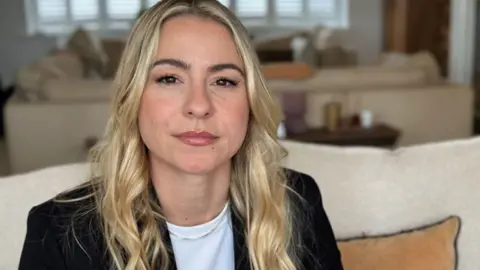 head and shoulders of Lucy Spraggan sitting on a cream sofa. She has long wavy blonde hair and is wearing a white tshirt and black blazer