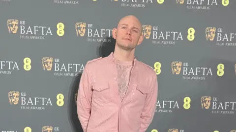 Jean Arnas Aidy Smith stands on a red carpet in front of a backdrop featuring repeated BAFTA Film Awards logos. He is wearing a light pink textured suit with matching trousers and white trainers, standing with hands behind his back.