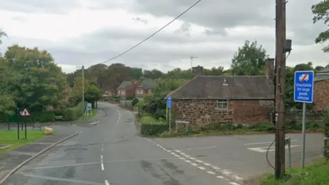 Google A road junction with a wooden pole in the foreground and a number of brown brick houses and trees on the far side